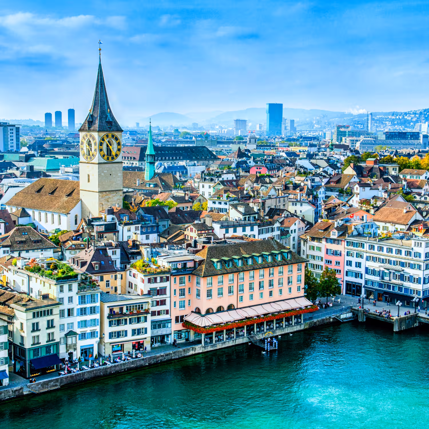 Aerial view of Zurich cityscape with the medieval St. Peter church clock tower and colorful riverside buildings along the Limmat River.