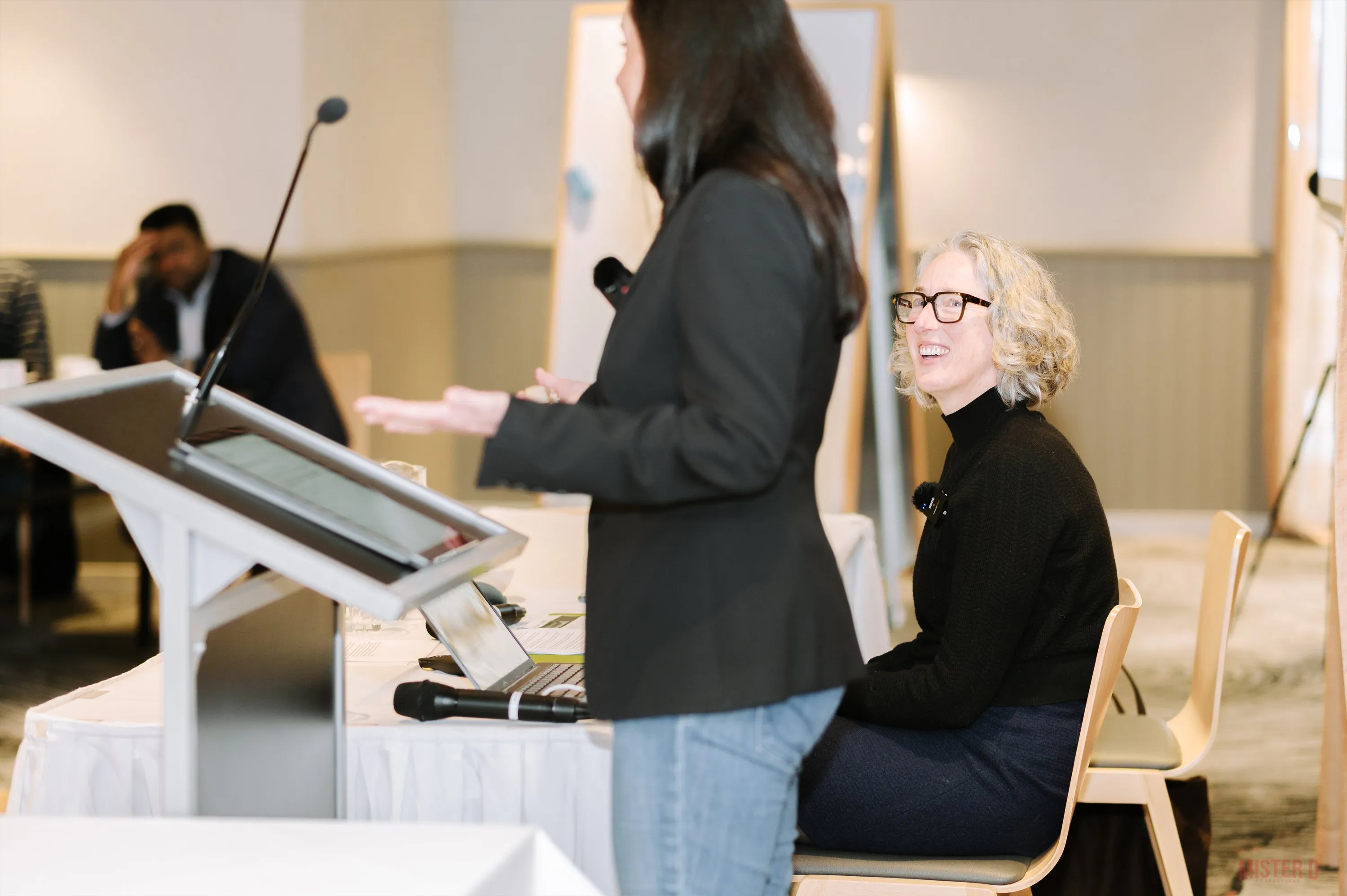 A podium speaker presenting to attendees, viewed from a different angle