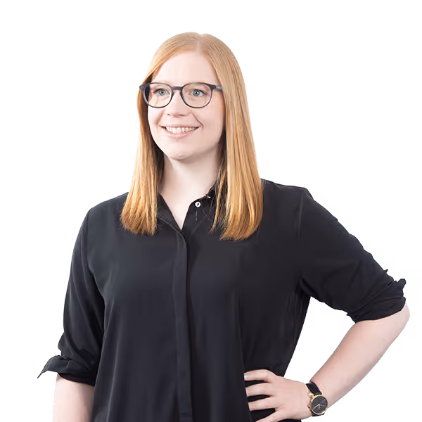 Frau mit roten Haaren und Brille lächelt, trägt ein schwarzes Hemd mit hochgekrempelten Ärmeln und eine Armbanduhr.