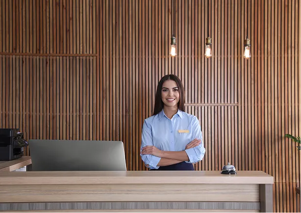 Smiling female receptionist standing behind front desk in a modern lobby with wooden slat wall and hanging light bulbs.