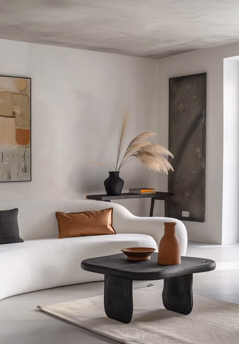Minimalist living room with a curved white sofa, black and brown cushions, black coffee table with terracotta vase and bowl, and a side table with black vase and dried pampas grass.