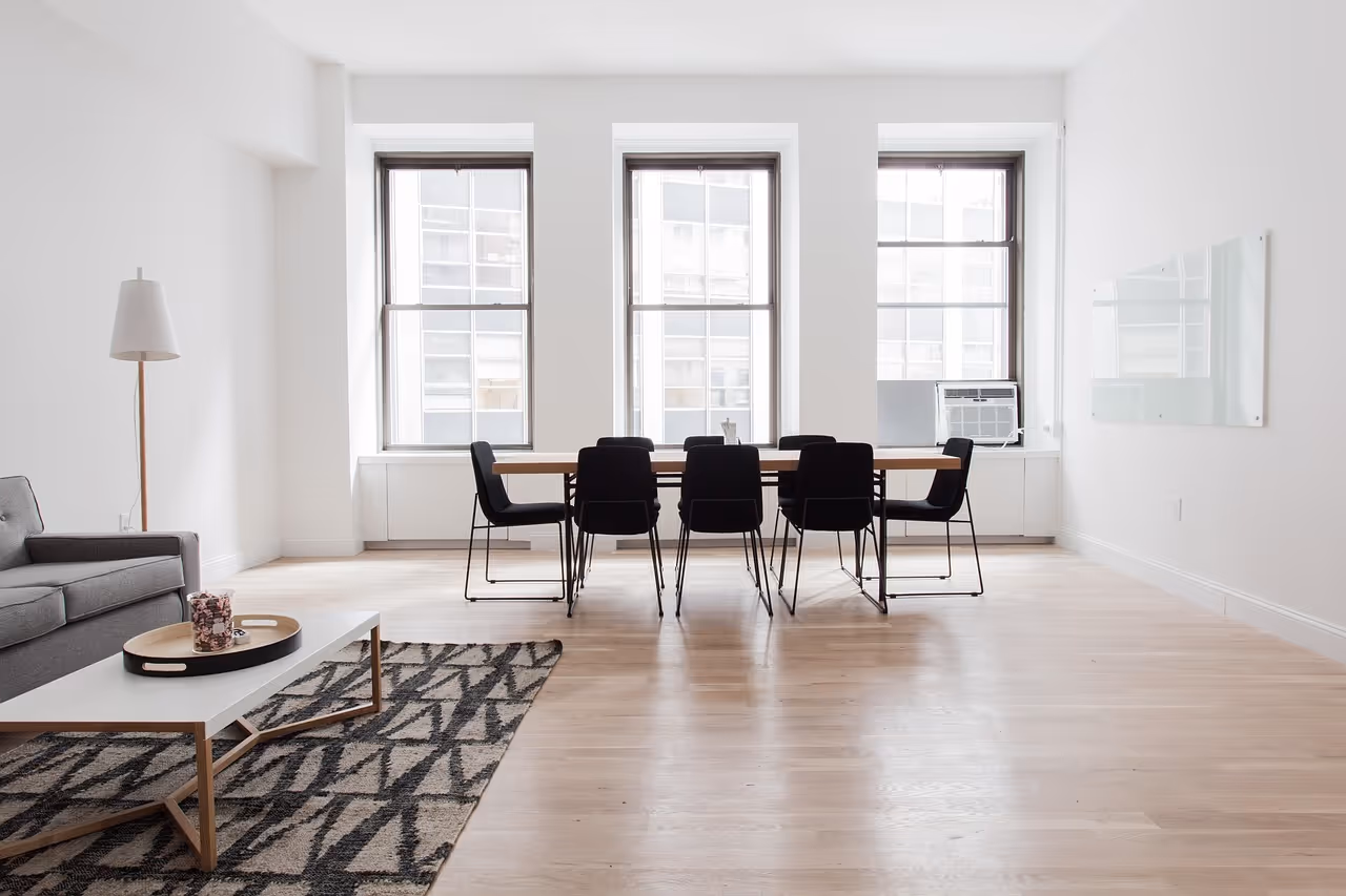 Minimalist office space with a large wooden table surrounded by black chairs, three tall windows, a gray sofa, patterned rug, and a floor lamp.