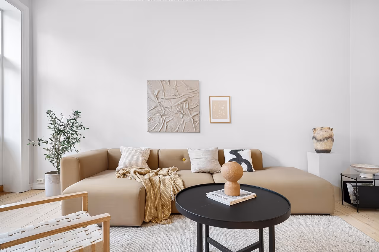 Modern living room with beige sectional sofa, three decorative pillows, a black round coffee table with a book and sculpture, and minimalist wall art.