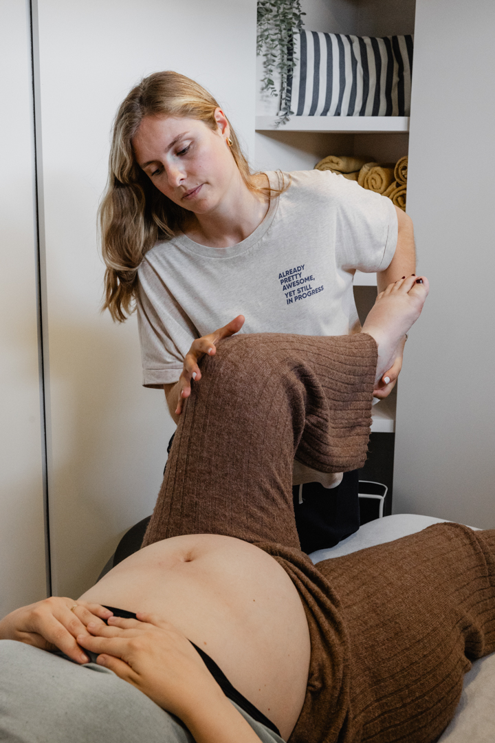 Een therapeut masseert voorzichtig het gebogen been van een zwangere vrouw op een massagetafel.