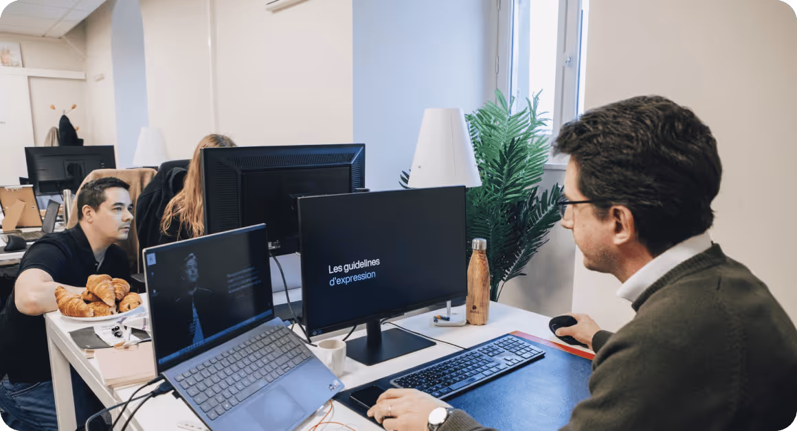 Deux hommes travaillent sur des ordinateurs dans un bureau, un écran affiche "Les guidelines d'expression".