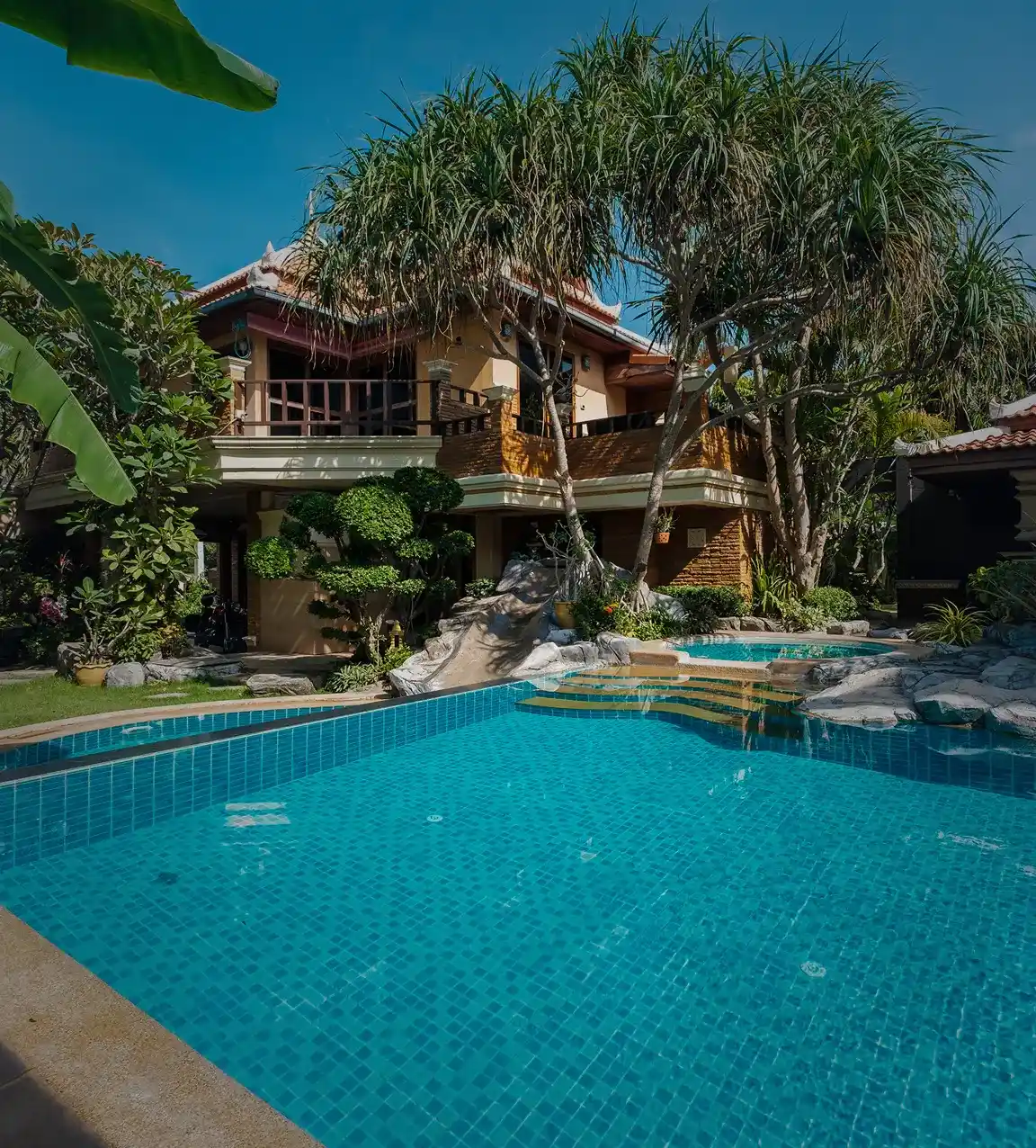 A stunning resort-style property with a clear blue swimming pool in the foreground, featuring natural rock accents and a small slide. In the background, a beautiful multi-story building with traditional architectural elements is surrounded by lush, tropical vegetation, including distinctive pandanus trees.