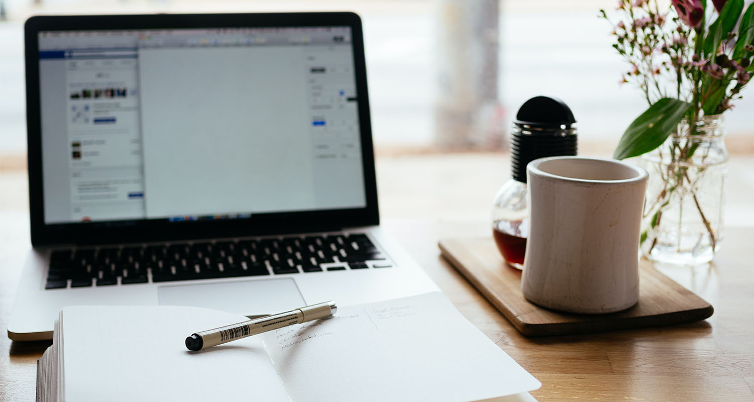 A desk setup with a notebook and pen, laptop and mug