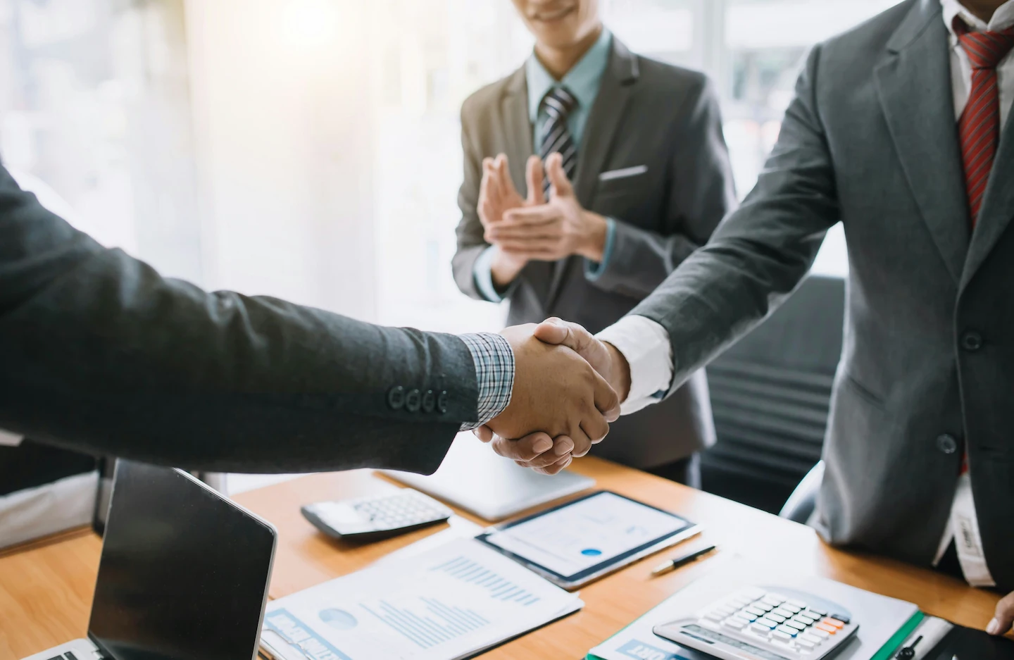 Two people shake hands across a desk on which various evaluation documents lie.