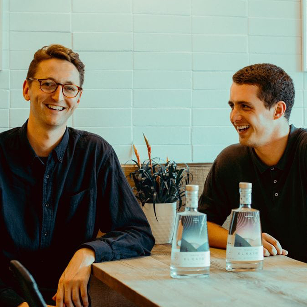 El Rayo Founders: Two laughing men leaning on a wooden table, with two bottles of tequila in the foreground. 