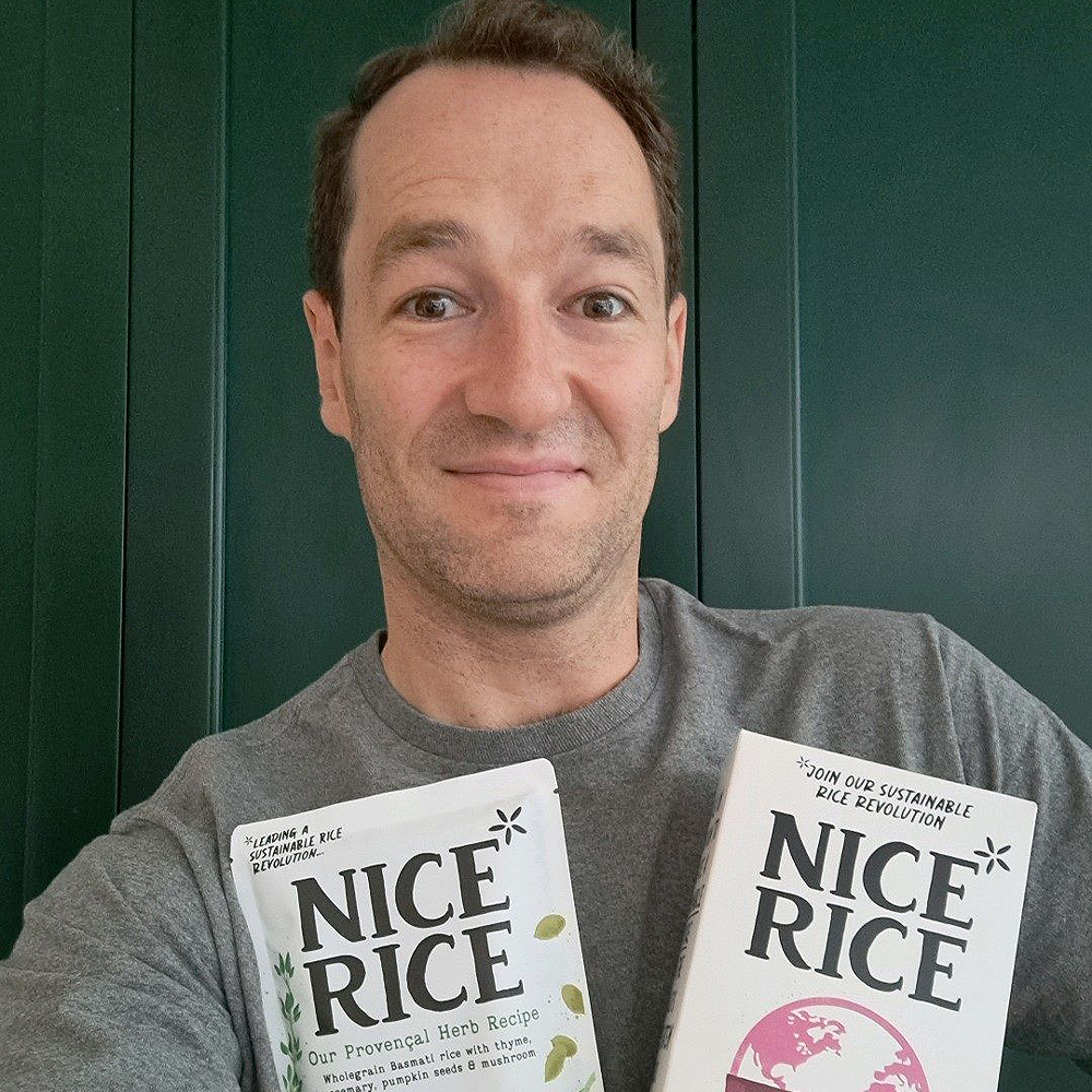 Nice Rice Founder: A man in a grey t-shirt taking a selfie with two packages of Nice Rice. 