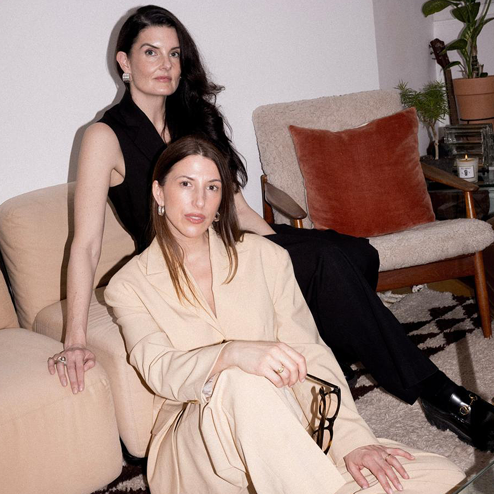 Nimbi Founders: Two women in suits with brown hair sat in a living room making eye contact with the camera. 