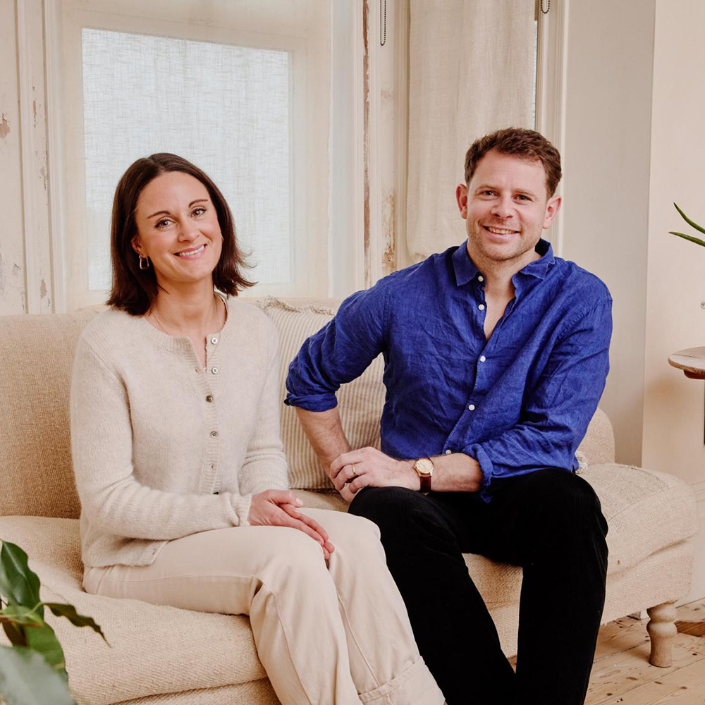 Naked Paper Founders: A man in a blue shirt sitting next to a woman with brown hair and white cardigan on a beige sofa. 