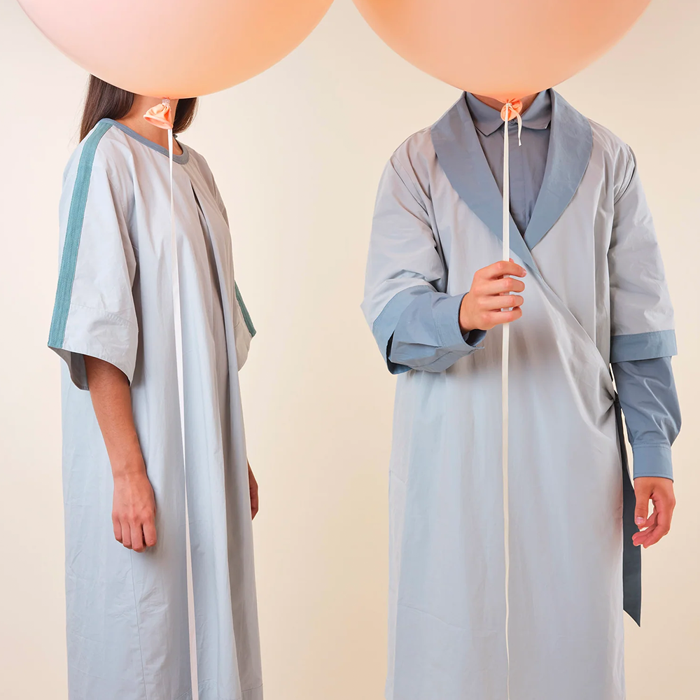 Two people in light blue hospital gowns, holding pink balloons over their faces. 