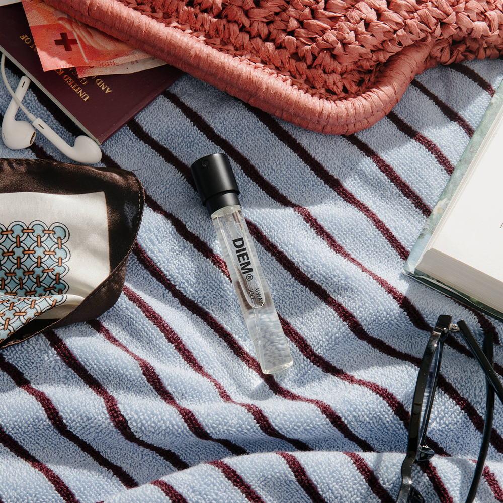 A perfume sample lying centre to a flat lay on a striped beach towel, with miscellaneous bag contents around the edges. 