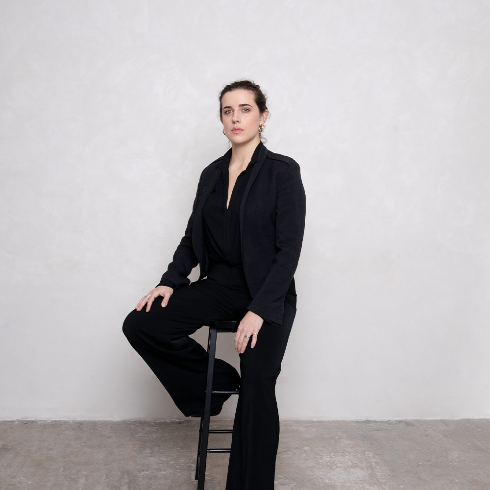 A picture of a woman in a black suit leaning against a stool on a white background.