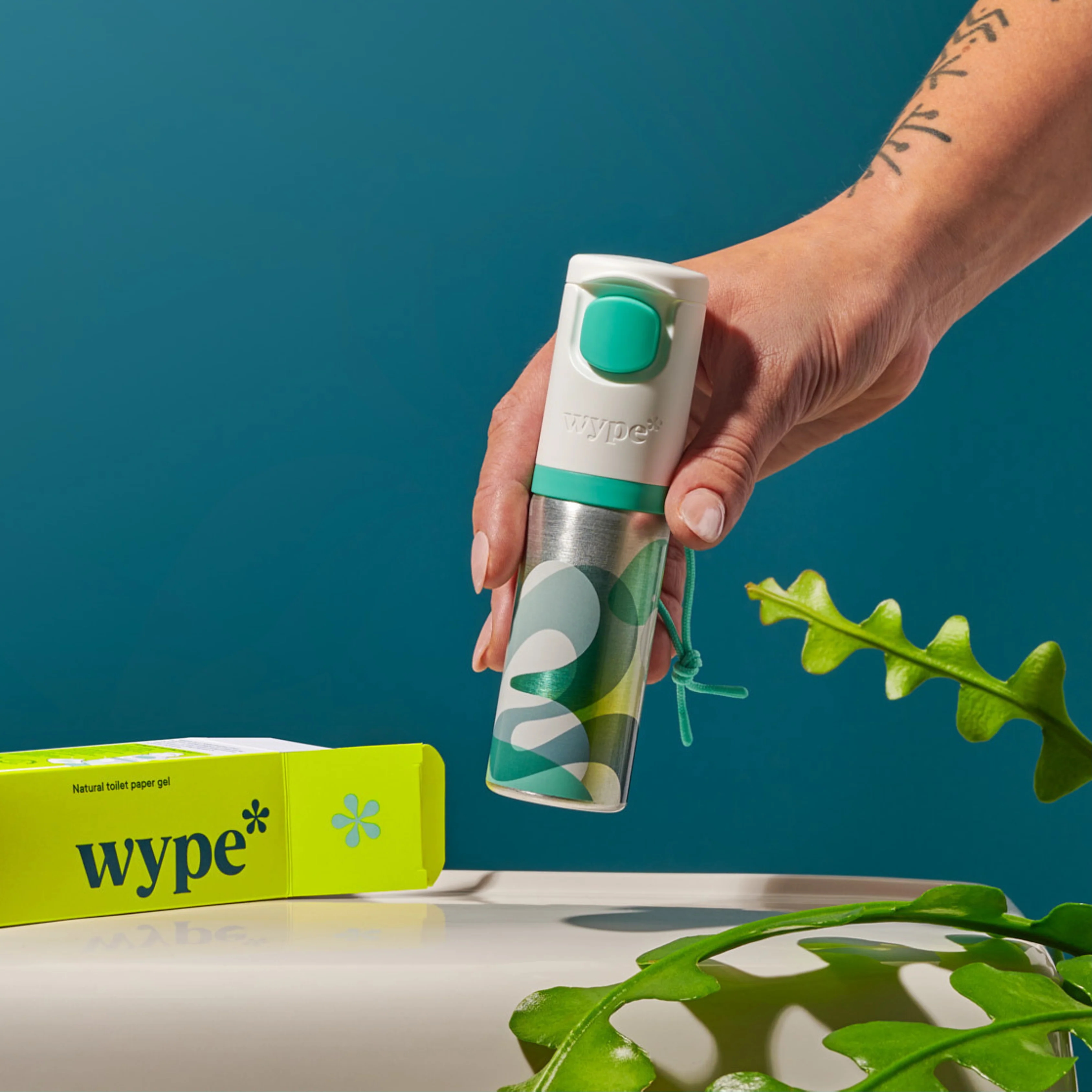 A hand holding a metallic Wype container. plants in the foreground and packaging box in the background.