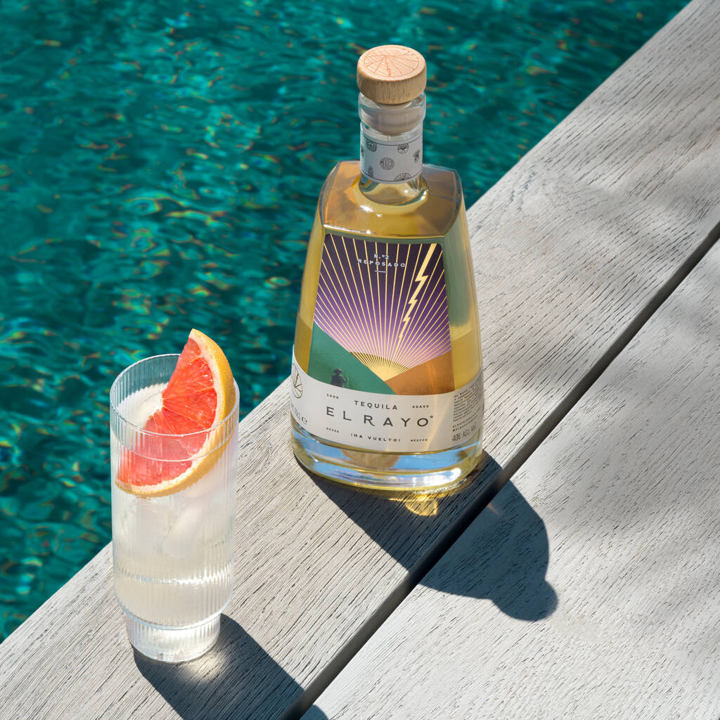 Bottle of tequila and glass with grapefruit slice positioned on the edge of a swimming pool.