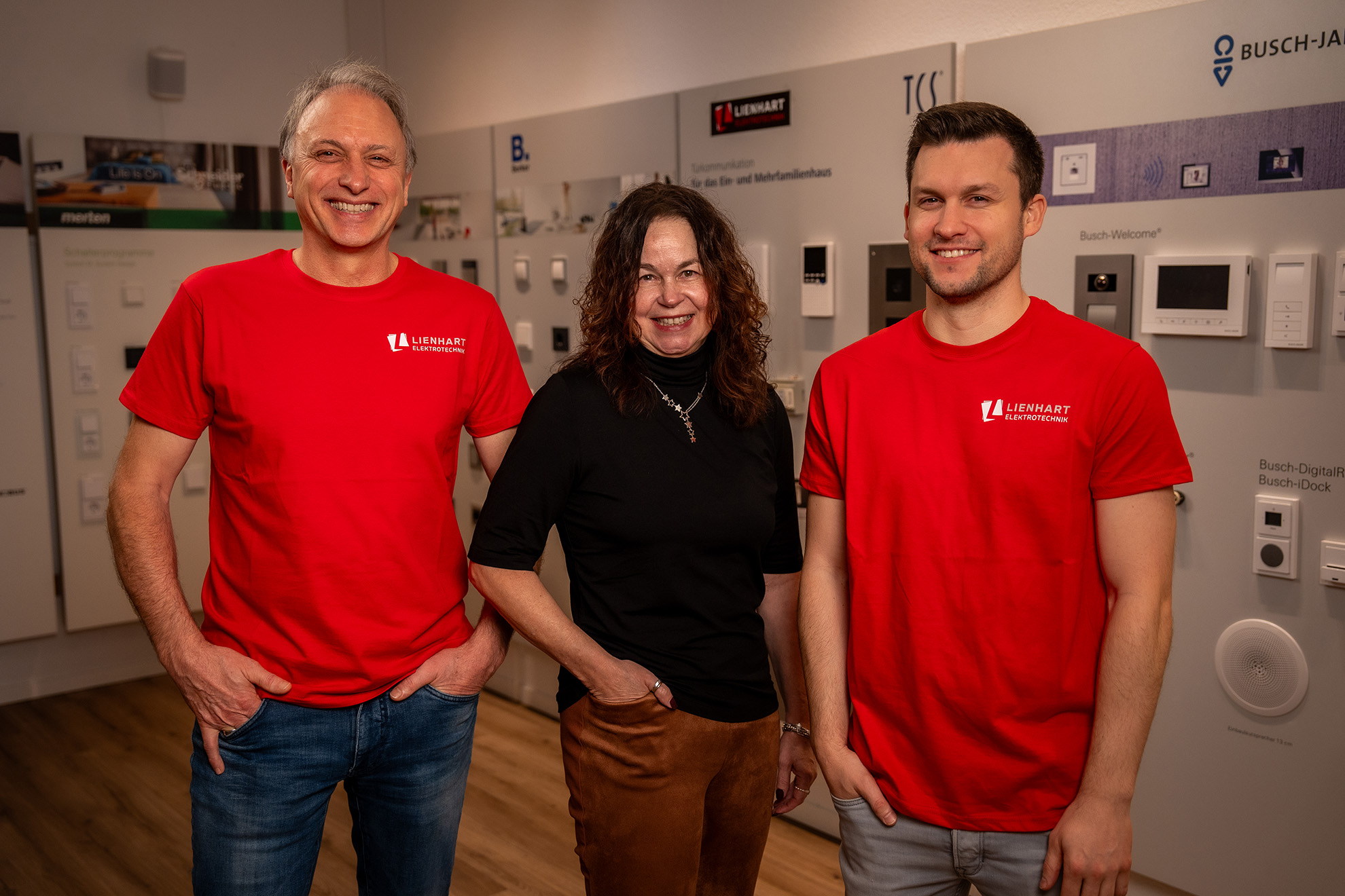 Drei lächelnde Personen stehen in einem Innenraum: zwei Männer in roten Lienhart Elektrotechnik T-Shirts und eine Frau in einem schwarzen Oberteil und brauner Hose.