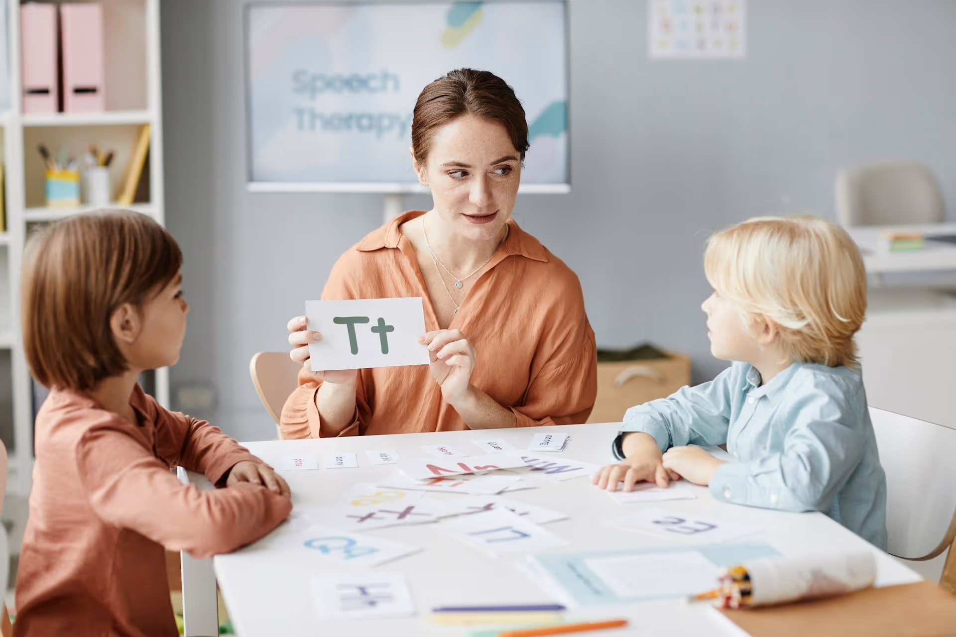Speech therapist holding a card with letter T while teaching two children seated at a table with alphabet cards.