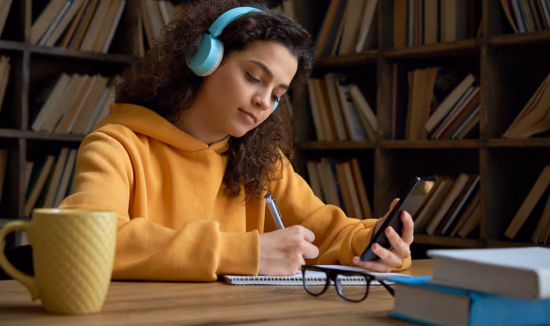 Young woman in a yellow hoodie wearing blue headphones, writing in a notebook while holding a smartphone, sitting at a table with books and a yellow mug.