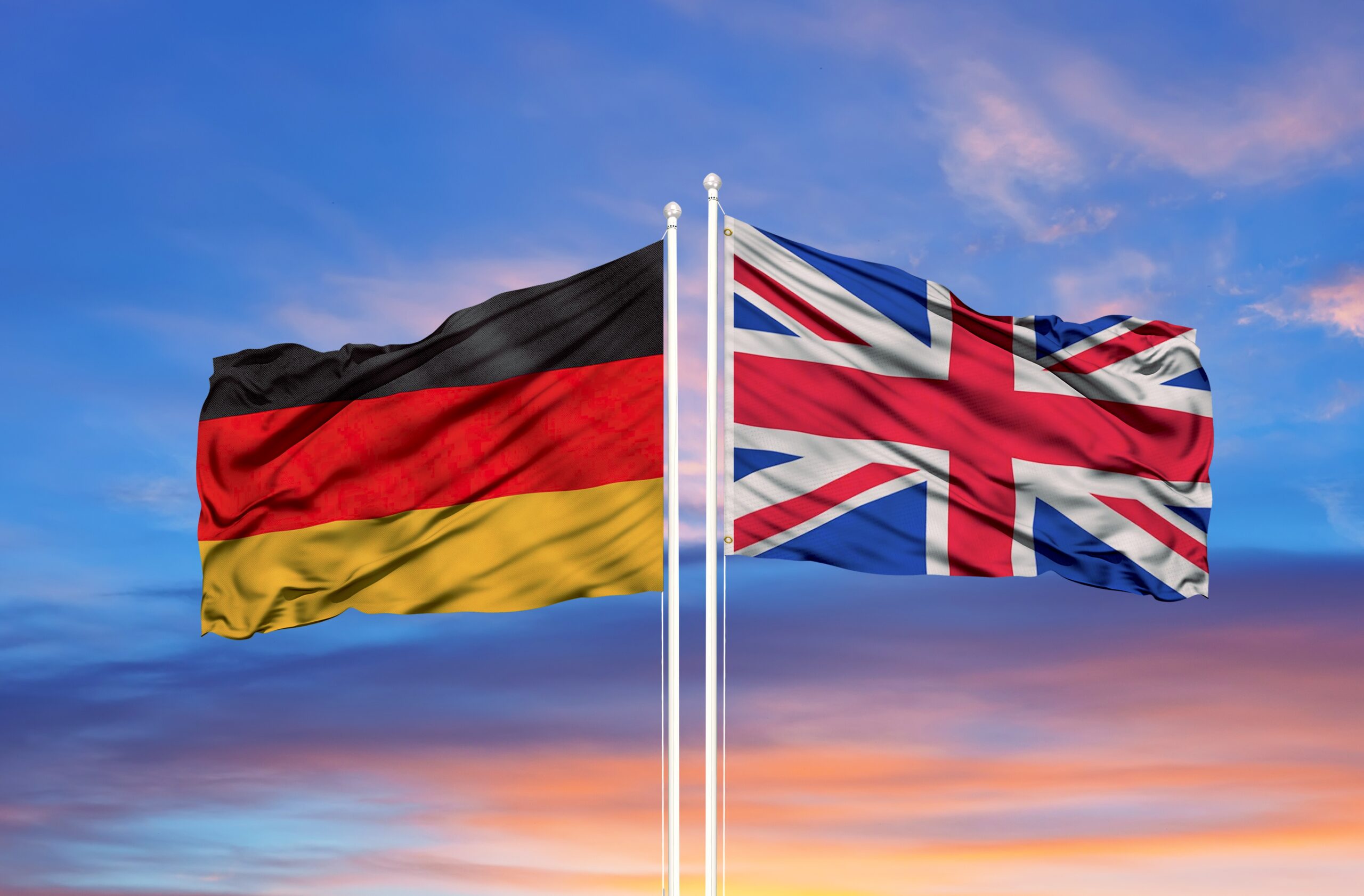 Flags of Germany and the United Kingdom waving on flagpoles against a colorful sunset sky.