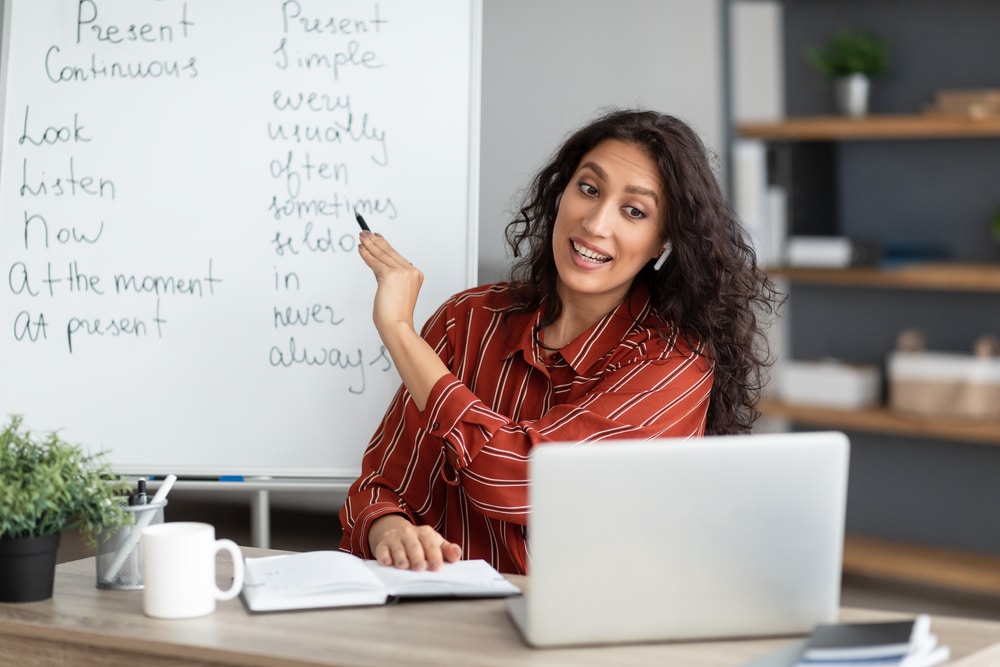 Woman teaching English grammar online, pointing at a whiteboard with present continuous and present simple tense examples.