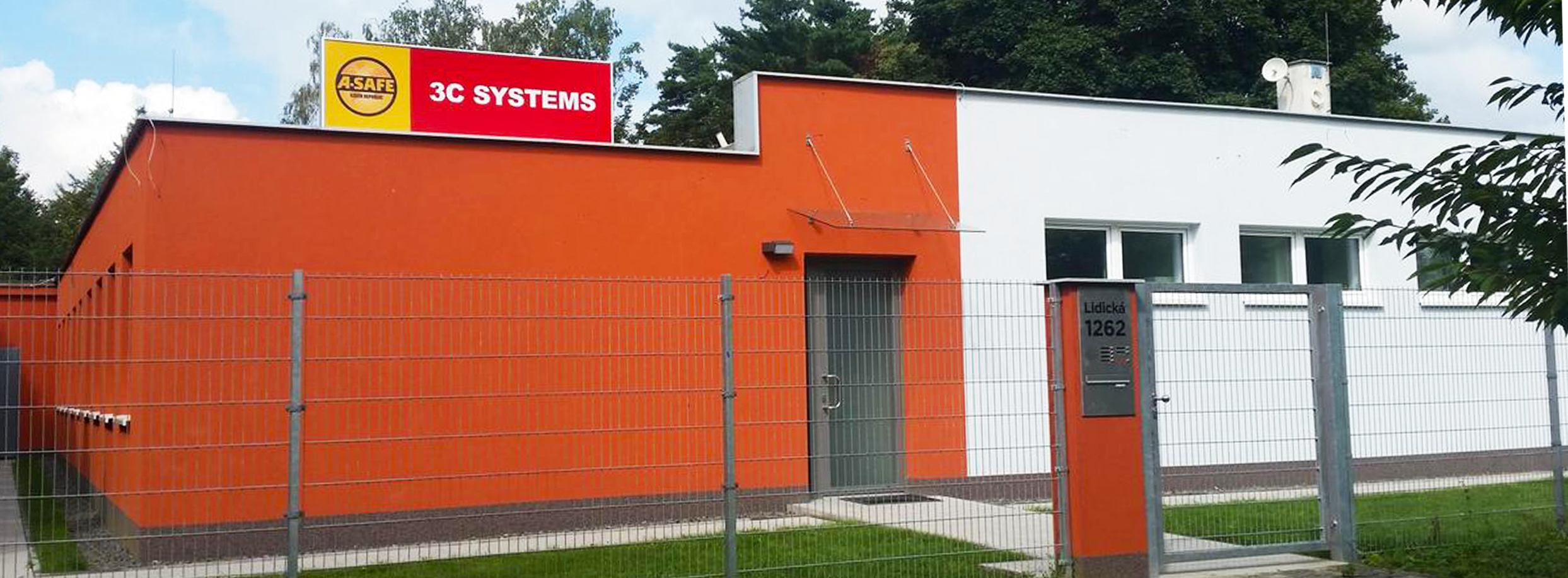 Budova s oranžovou a bílou fasádou s nápisem 3C SYSTEMS a ohradou s kovovou bránou.