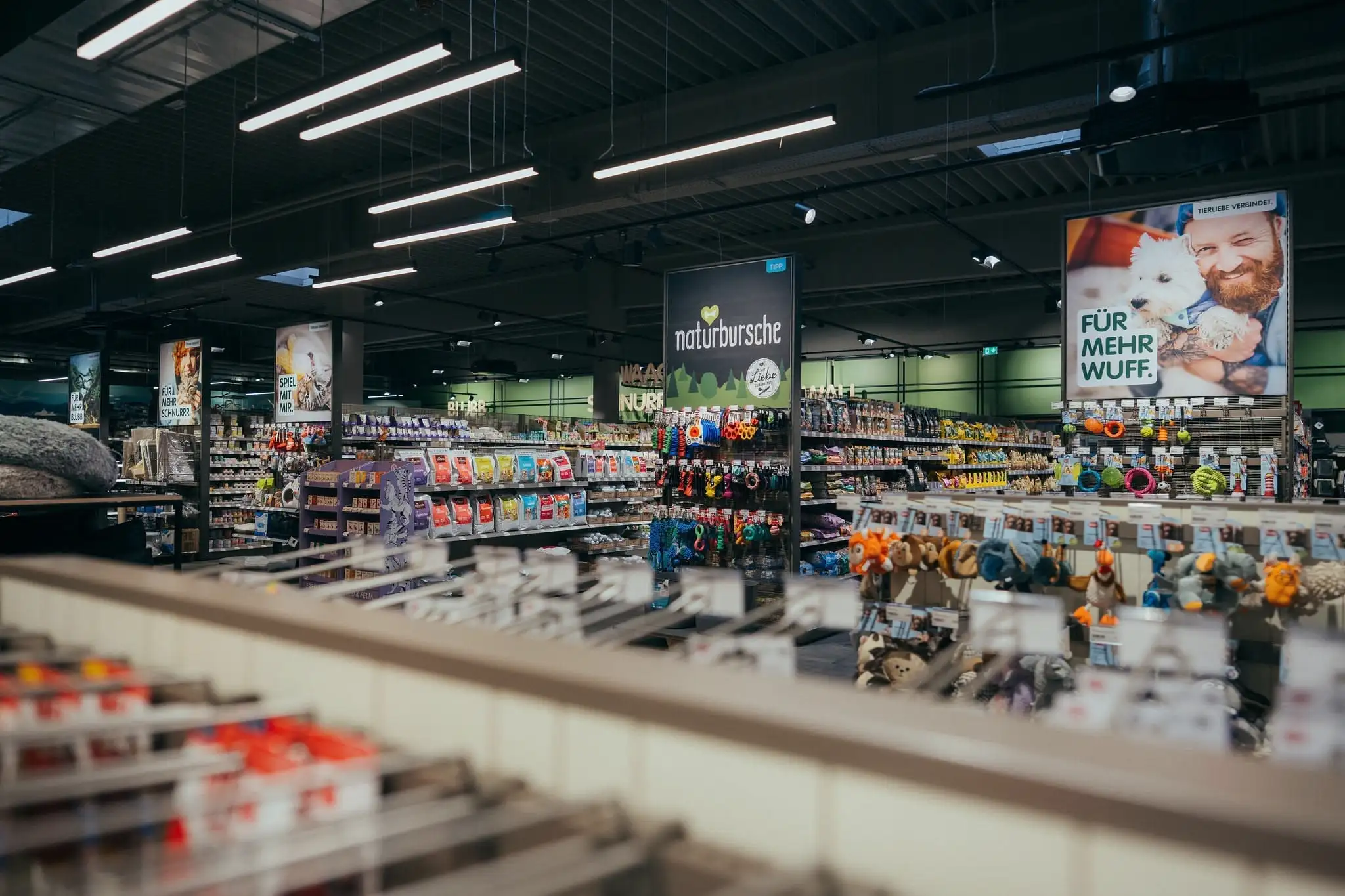 Interior view of a well-lit pet store with shelves stocked with pet food, toys, and accessories, featuring large signs and advertisements.