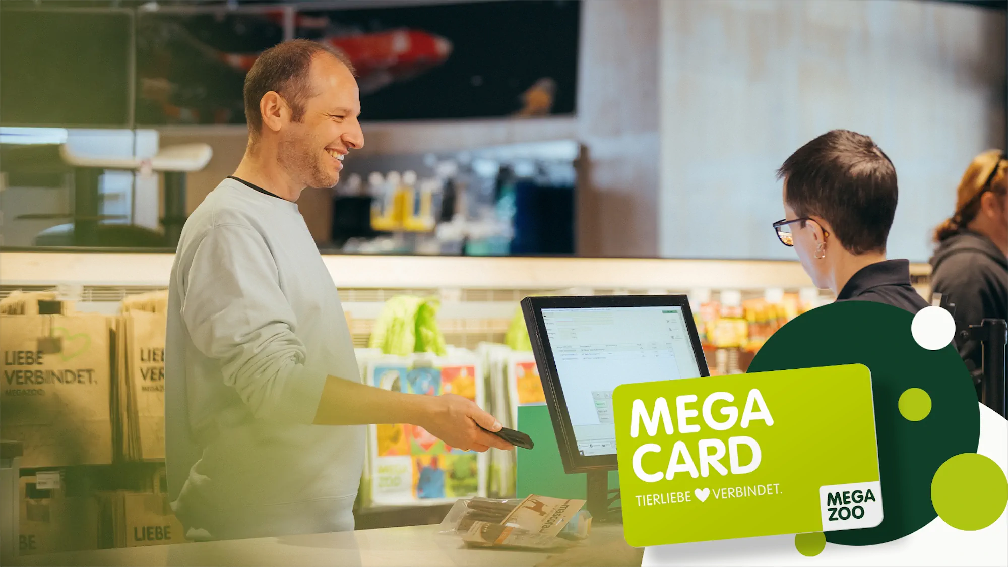 Smiling man paying at a checkout counter using a card device with a cashier in the background and Mega Card loyalty program branding overlay.