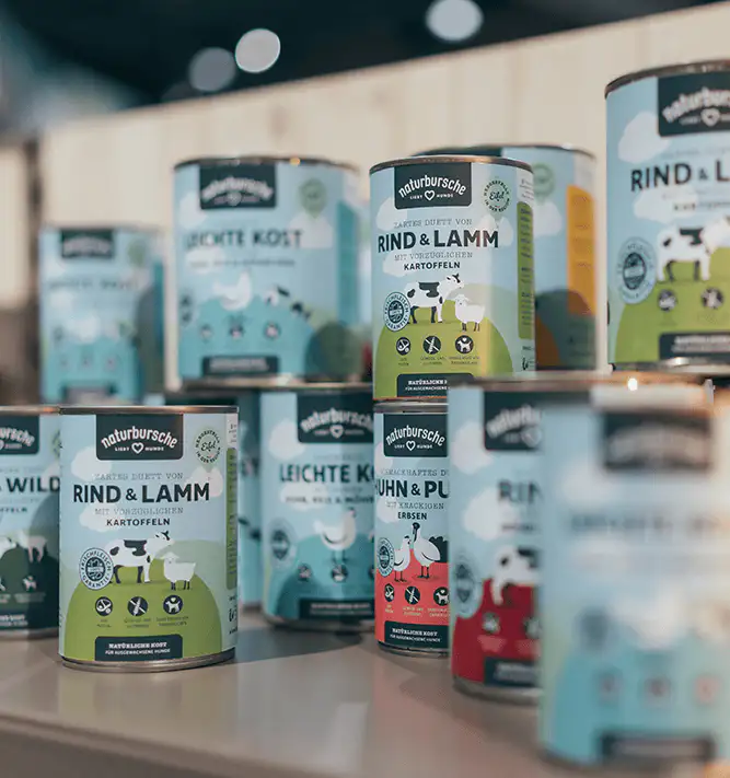 Cans of Naturbursche brand pet food with labels showing varieties like beef and lamb, chicken and peas, and light diet on a display table.
