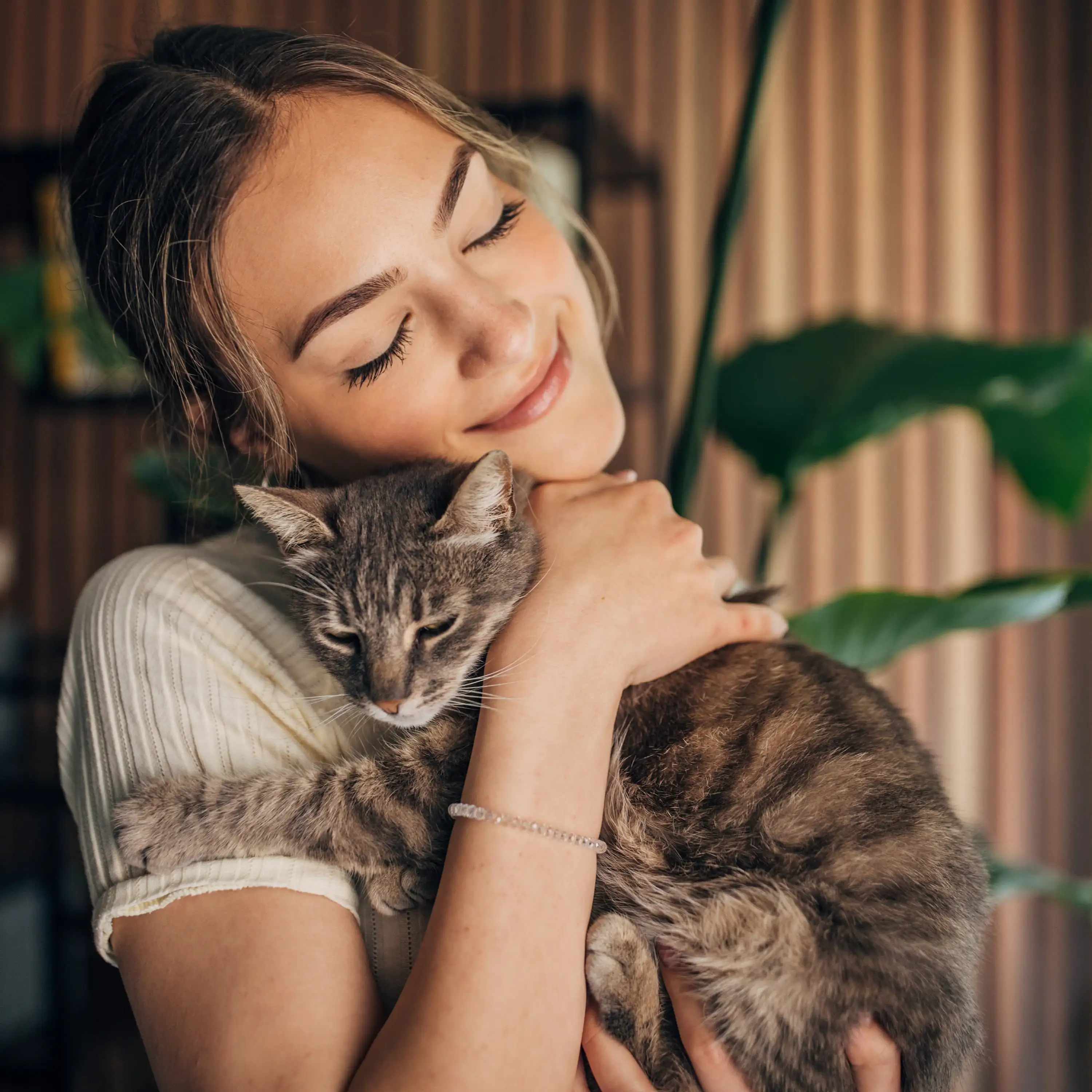 Woman with closed eyes smiling and hugging a gray tabby cat indoors.