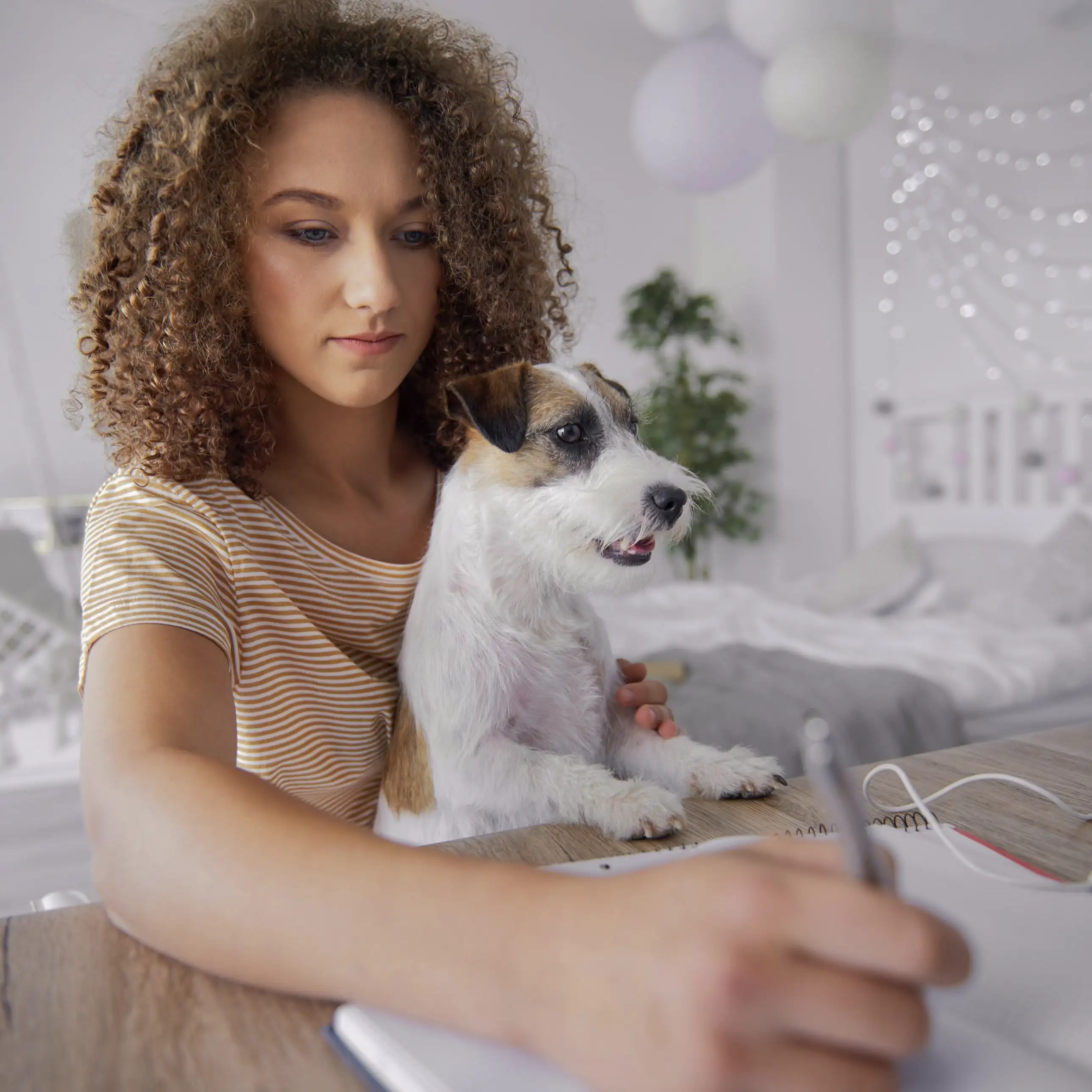 Woman with curly hair holding a small dog while writing at a desk in a cozy room.