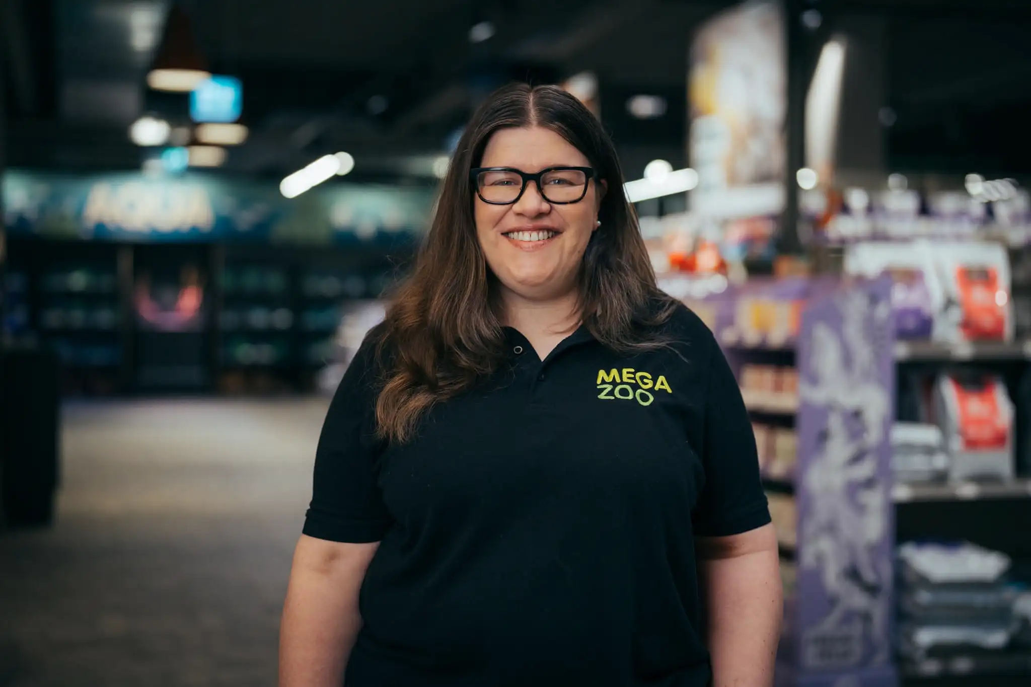 Smiling woman with long brown hair and glasses wearing a black Mega Zoo polo shirt inside a store.
