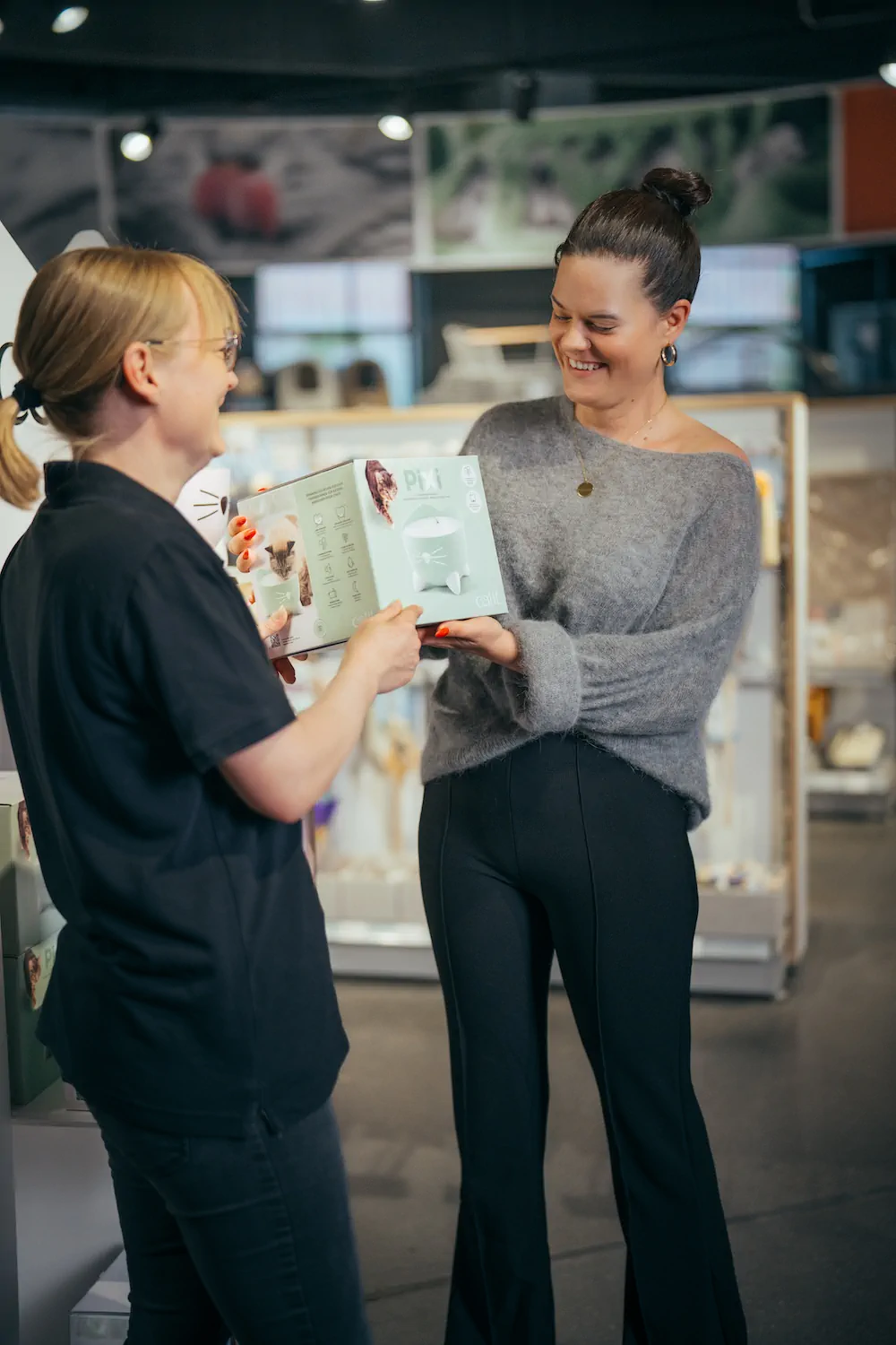Two women smiling and holding a pet product box in a store with pet supplies in the background.