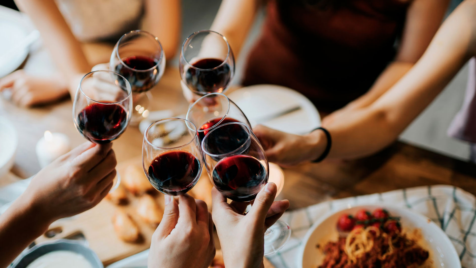 Group of people toasting with glasses of red wine around a dinner table, with plates of food in the background.