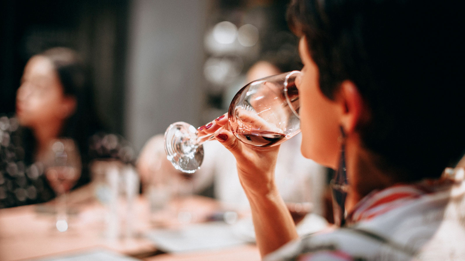 Close-up of a woman tasting red wine from a glass during a wine tasting session, with other participants blurred in the background.