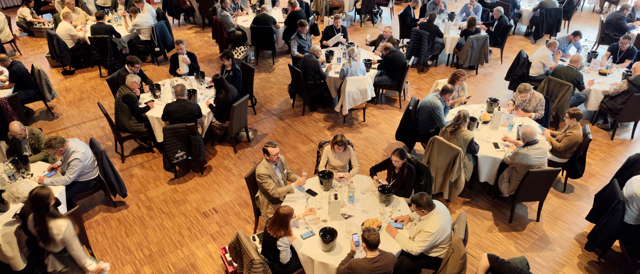 Overhead view of a competition tasting room, judges scoring wines from their own devices at round tables.