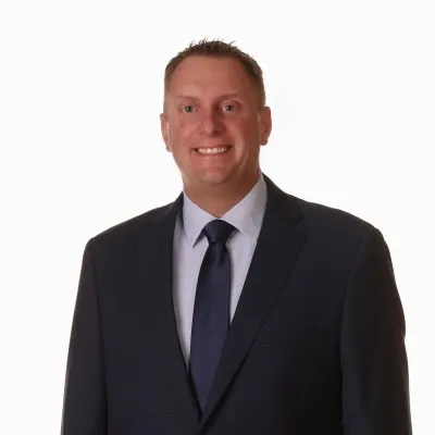 Smiling man wearing a dark suit, white shirt, and dark tie against a plain white background.