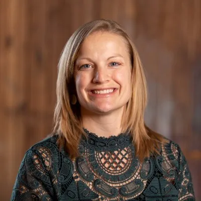 Smiling woman with shoulder-length blonde hair wearing a dark green lace top with a wood background.
