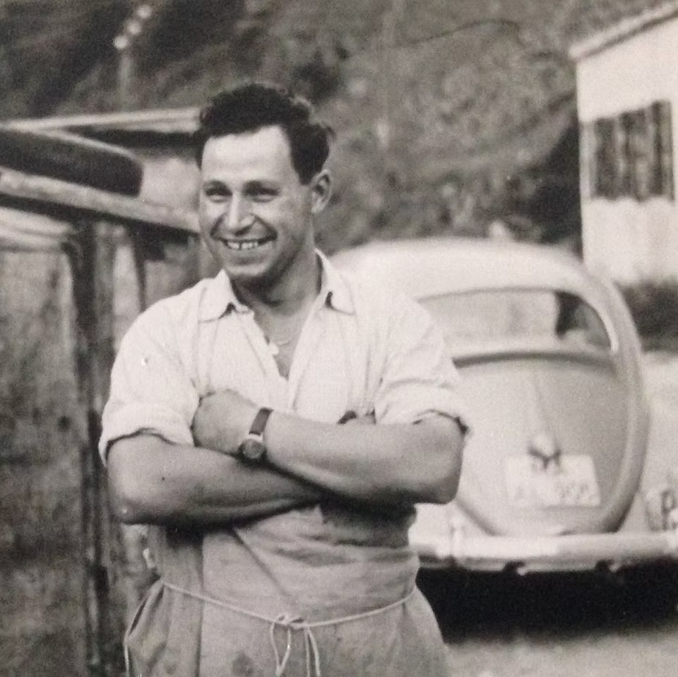 Schwarz-weiss-Foto von Peter Volgger, dem Gründer, wie er lächelnd mit verschränkten Armen in Arbeitskleidung vor einem Volkswagen Käfer steht; im Hintergrund ein Gebäude mit Fensterläden.
