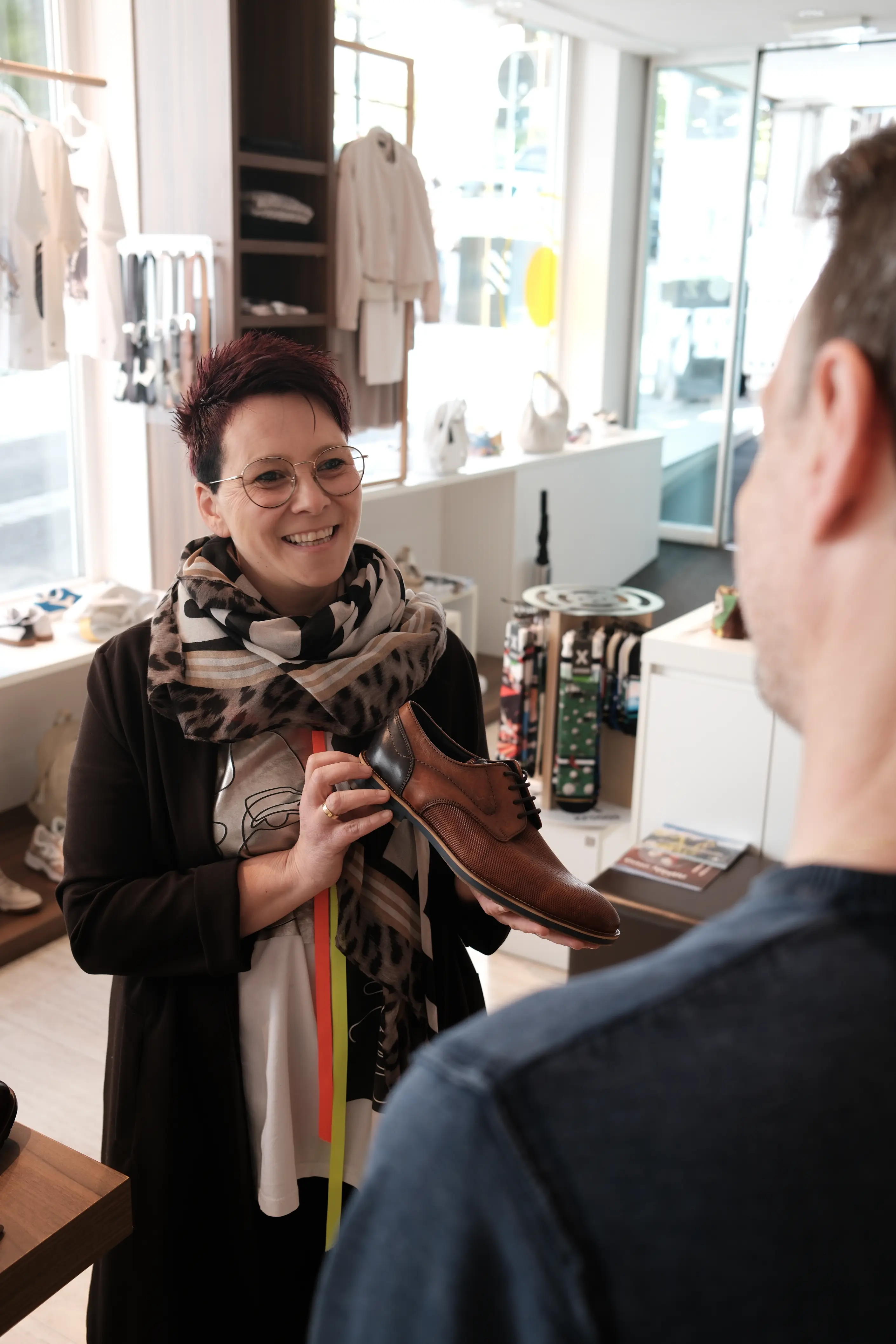 Verkäuferin in einem modernen Schuhgeschäft zeigt einem Kunden lächelnd einen braun-schwarzen Lederschuh; im Hintergrund sind Regale mit Kleidung und Accessoires sichtbar.
