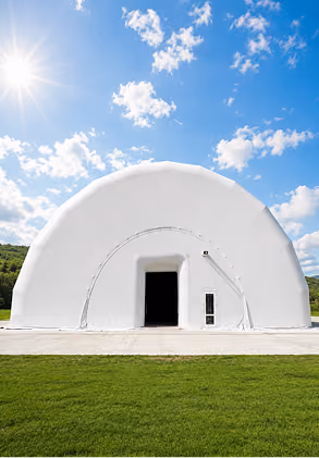 White dome-shaped building with an open doorway, grass in front, and blue sky with clouds and sun.