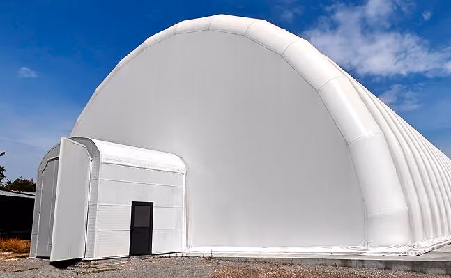 Large white inflatable dome structure with a small attached entrance building under a partly cloudy sky.