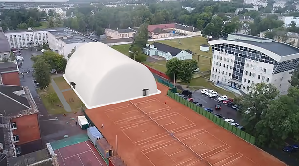 Aerial view of red clay tennis courts next to a white dome building and surrounding urban area.