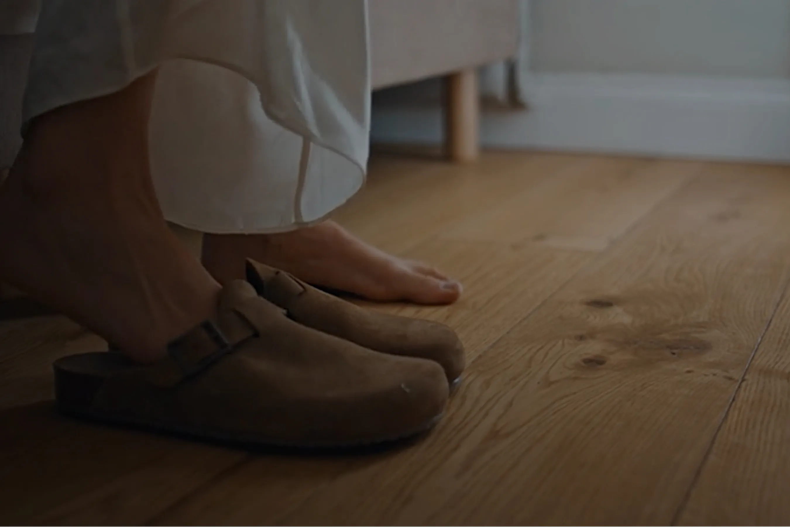 An image that shows a lady waking up from bed and putting her slippers on. You can only see the feet at the edge of the bed with one foot slding into the right slipper.