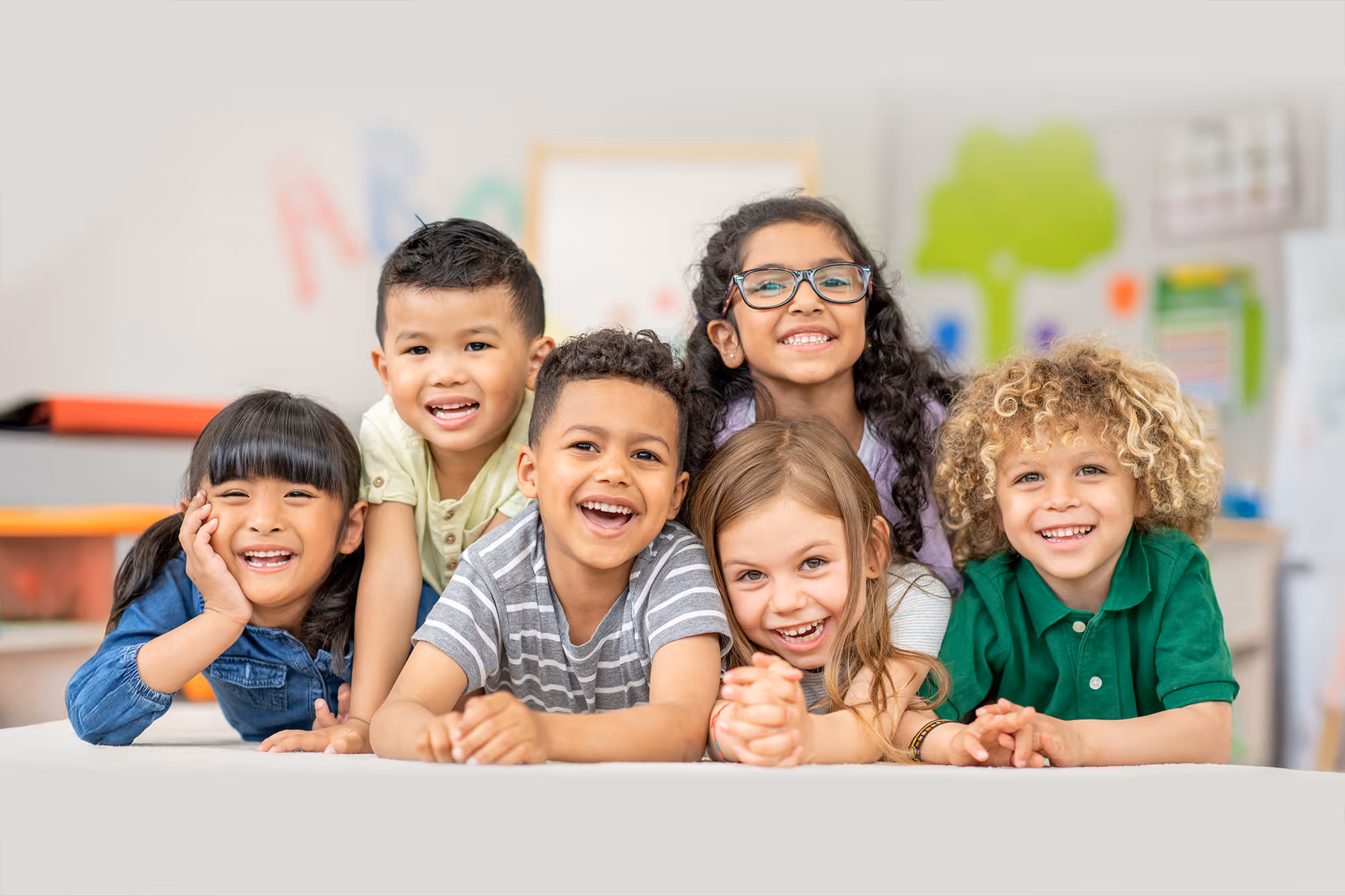 Six diverse children lying on their stomachs, smiling and looking at the camera in a bright classroom.