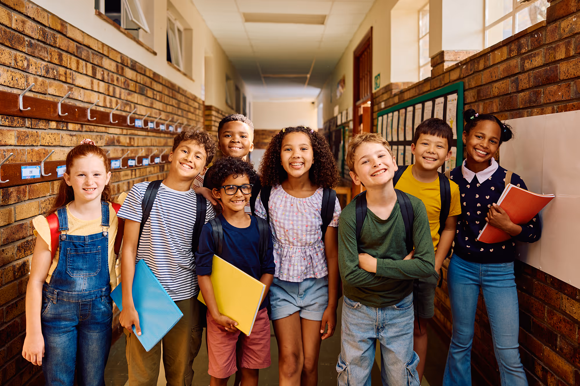 A group of eight smiling diverse children standing together in a school hallway, holding folders and backpacks.