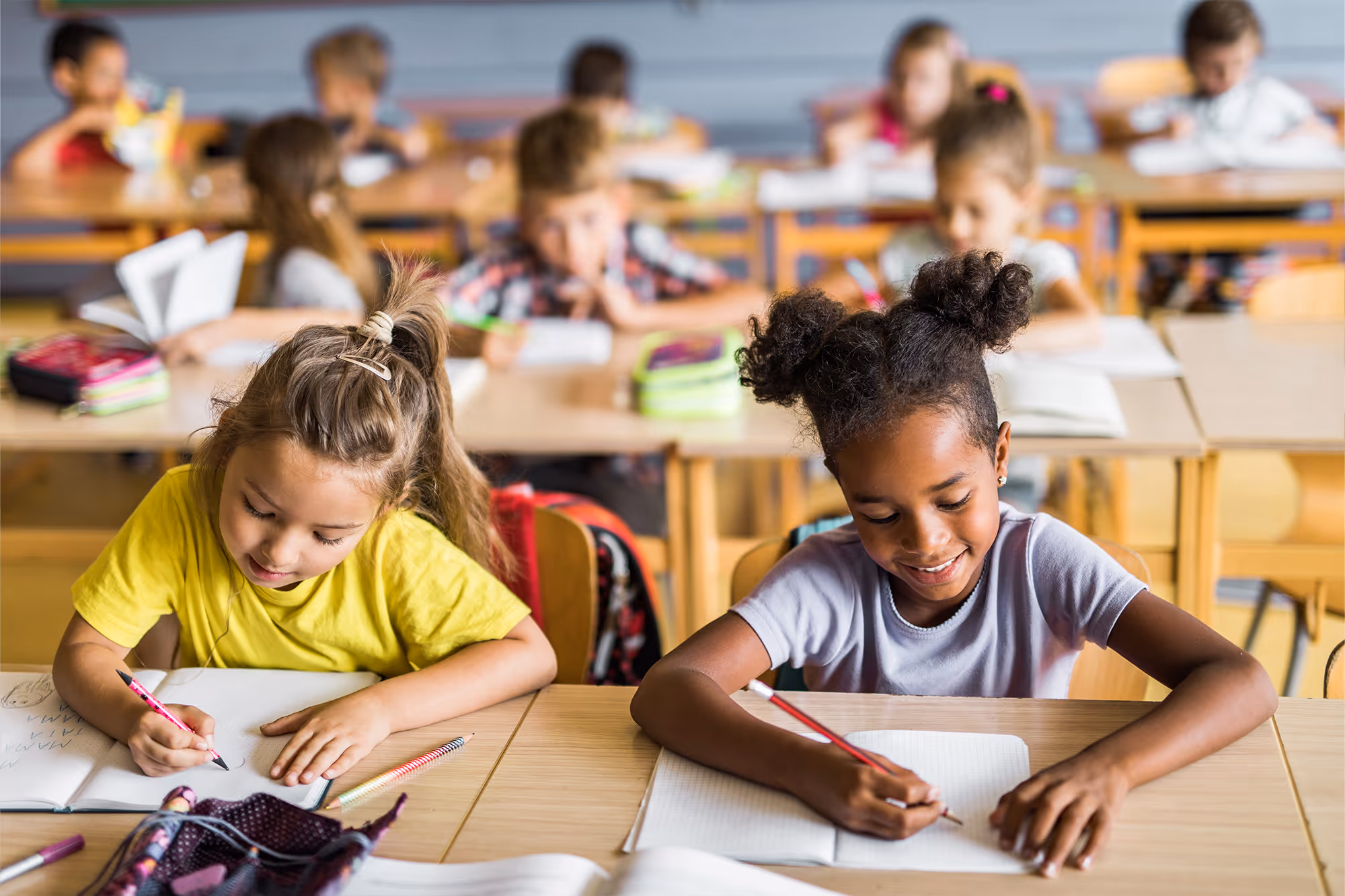 Two young girls smiling and writing in notebooks at a classroom desk with other children blurred in the background.