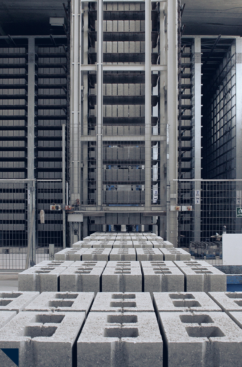 Rows of stacked concrete blocks in an industrial warehouse with machinery and metal shelving.