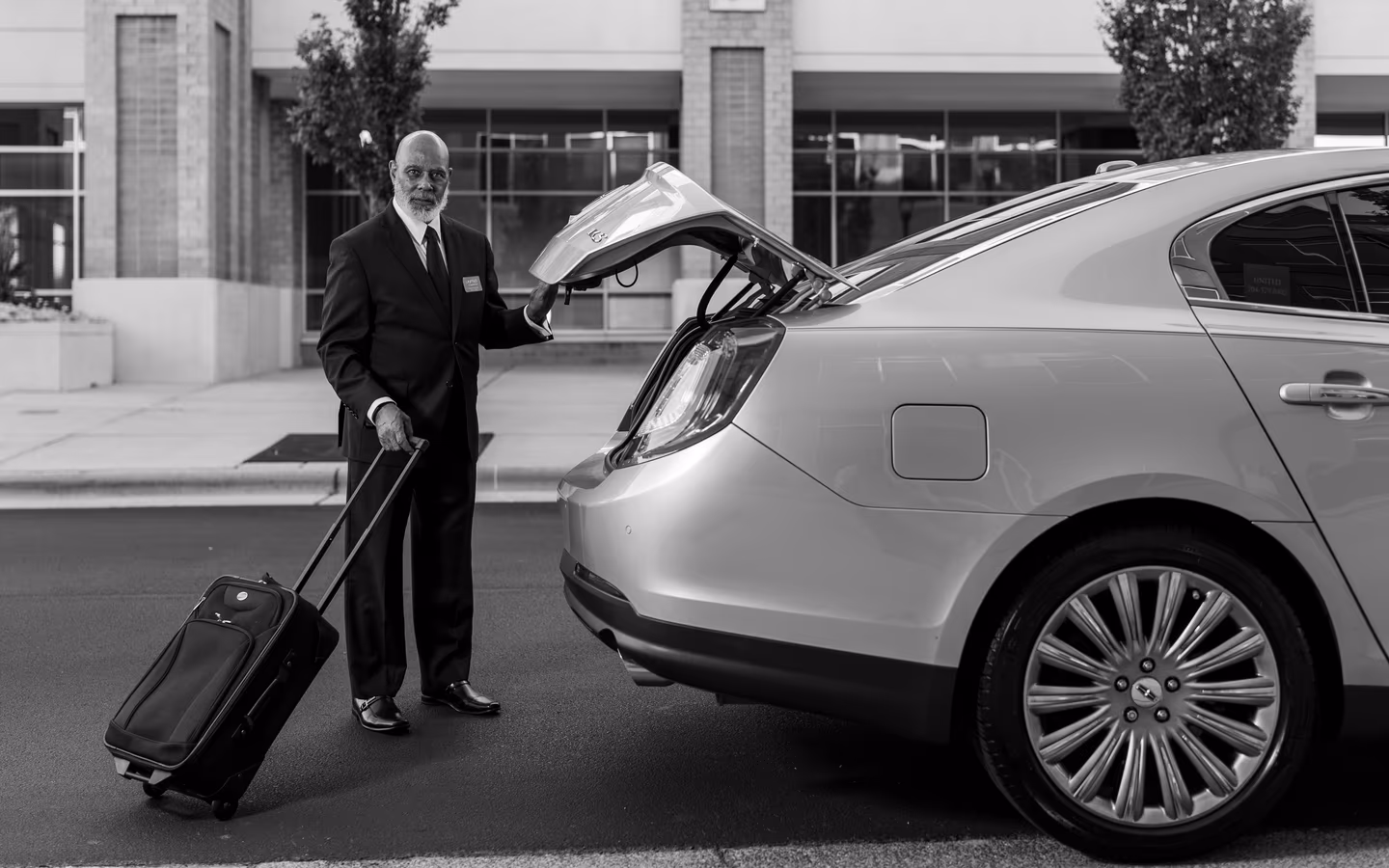 Chauffeur in suit loading luggage into luxury car trunk outside a modern building.